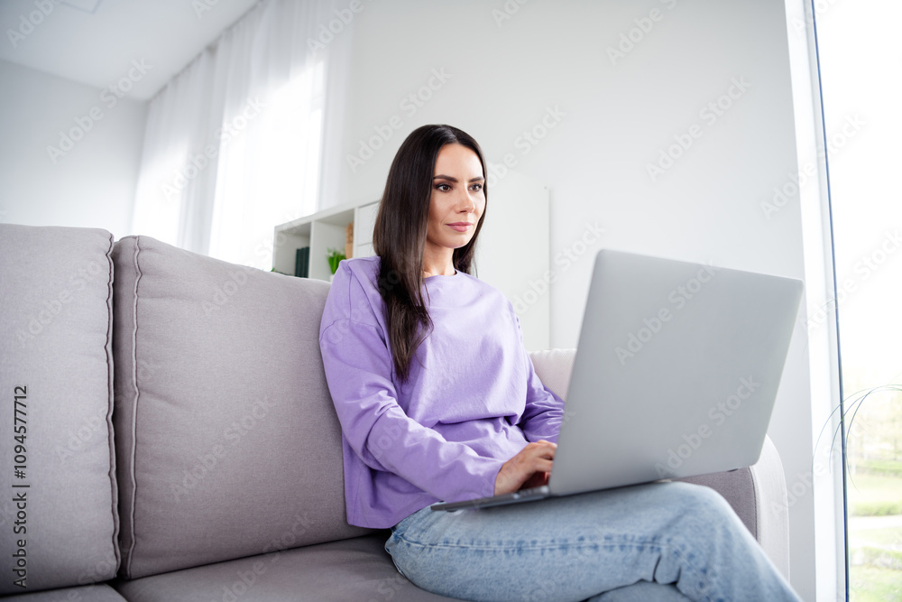 Naklejka premium Young woman enjoying a peaceful weekend at home, lounging on the sofa while working on her laptop in a bright living room.