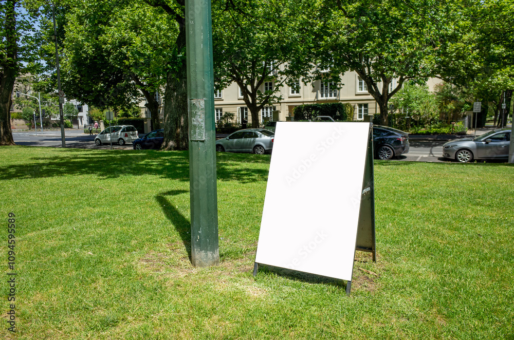 Background texture of white, blank A-frame mockup sandwich board sign stands on a grassy median or outdoor green space in an urban park. Public notice announcement display. Copy space for design. Background texture of white, blank A-frame mockup sandwich board sign stands on a grassy median or outdoor green space in an urban park. Public notice announcement display. Copy space for design.