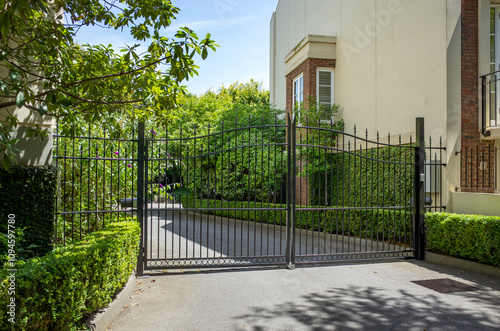 A wrought iron security gate guards the entrance to an upscale property with well-maintained greenery. A gated residential complex with a locked entrance. photo