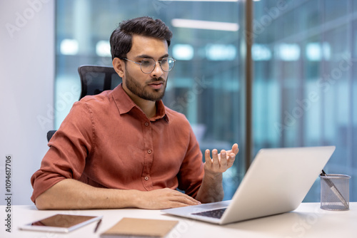 Latin American man businessman participating in online video call on laptop, seated in modern office with casual attire. Engaging in virtual meeting, expressing communication and professionalism.