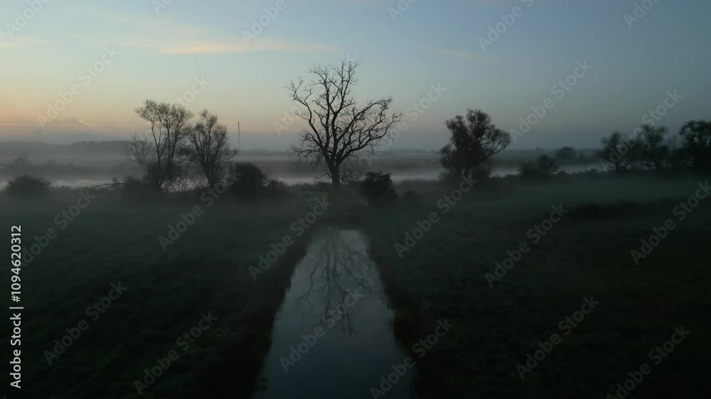 A foggy sunrise over a quiet marshland with a still stream and bare tree reflections A foggy sunrise over a quiet marshland with a still stream and bare tree reflections