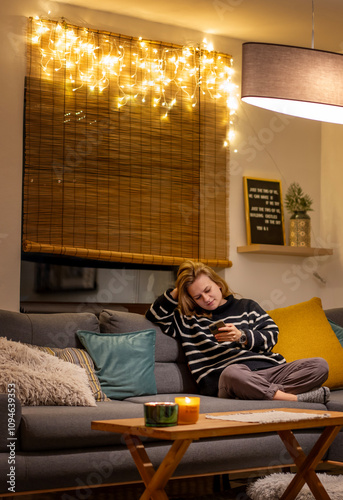 Young woman spends a cozy winter evening at home sitiing on the sofa looking at her smartphone. Winter holidays, Christmas and online surfing concept photo