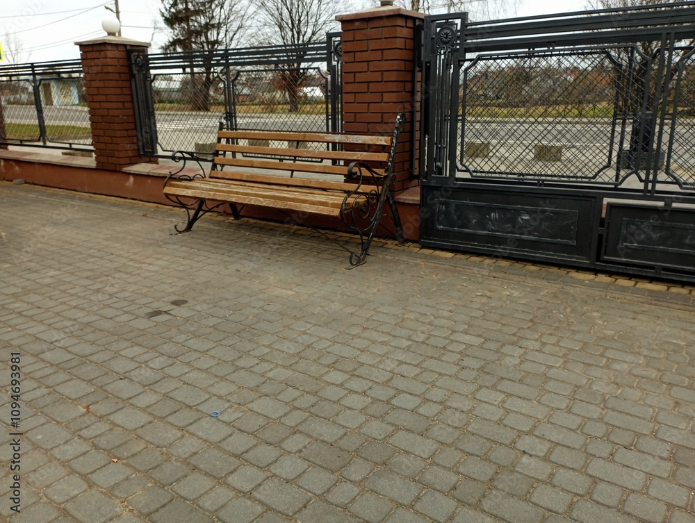 Wooden bench near the fence in the yard of the house. Bench for relaxation in the yard. The yard is paved with paving stones and is fenced with a metal fence. Wooden bench near the fence in the yard of the house. Bench for relaxation in the yard. The yard is paved with paving stones and is fenced with a metal fence.