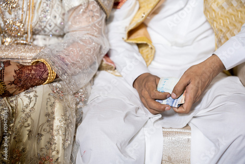 Pakistani groom holding one thousand rupee bank note in his hand wearing white Shalwar Qameez and golden waist coat with bride. photo