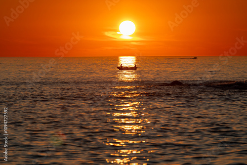 A serene sunset captures the vibrant orange sky and vast ocean, while a silhouetted boat floats gently, creating a peaceful and calming scene on the horizon. photo