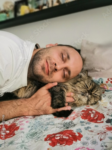 A man is sleeping, cuddling a long-haired cat on a bed with a colorful floral blanket. The cozy scene captures love for pets and emotional connection. Great for family and home themes.