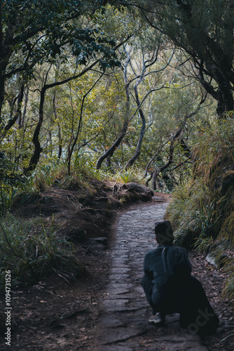 Woman crouched in the middle of a trail, watching a cat in the background in a levada, Madeira Portugal