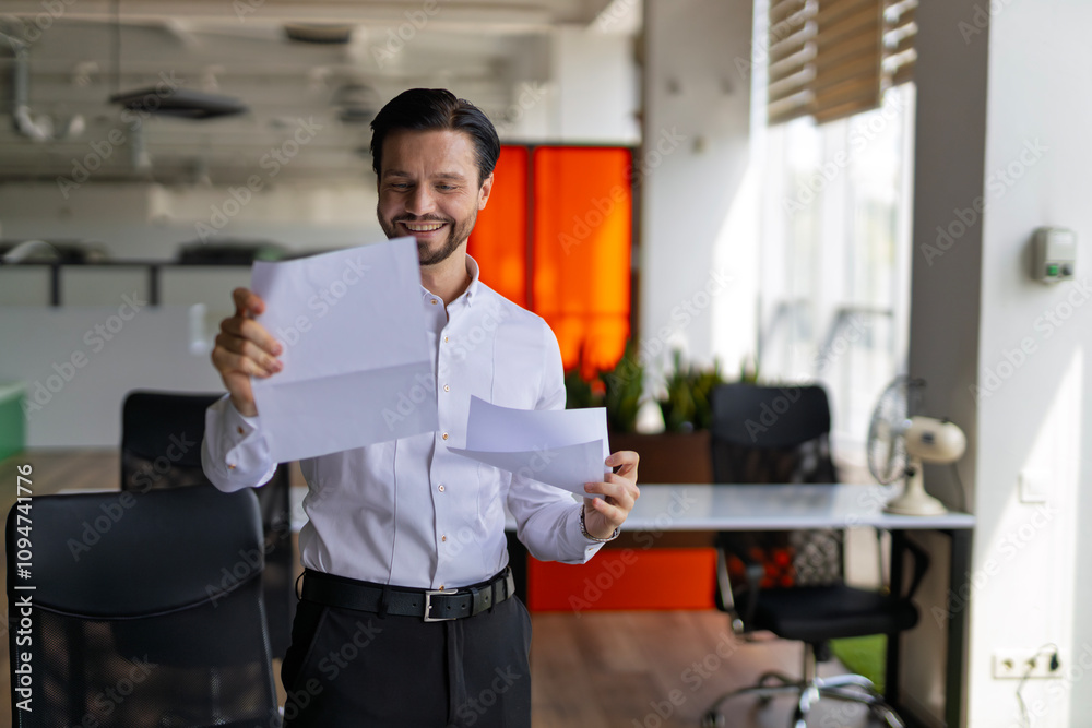 A man is standing in a room with a white paper in his hand. He is smiling and looking at the paper. The room has a modern design with a few chairs and a potted plant A man is standing in a room with a white paper in his hand. He is smiling and looking at the paper. The room has a modern design with a few chairs and a potted plant