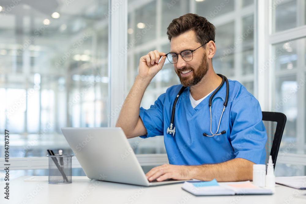 Confident man doctor in blue scrubs working on laptop in bright office, adjusting glasses, showing professionalism. Modern healthcare setting with medical tools on desk.
