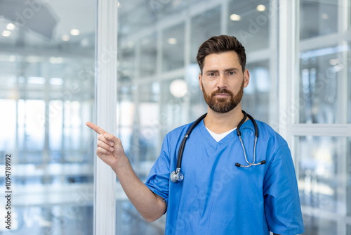 Confident man doctor in blue scrubs standing indoors at modern hospital pointing to side. Professional with stethoscope demonstrates healthcare guidance, authority, and trust in medical field.