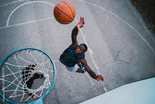 A basketball player attempts a dunk under the hoop on a concrete court, viewed from above, highlighting power, athleticism, and precision in an outdoor sports setting.