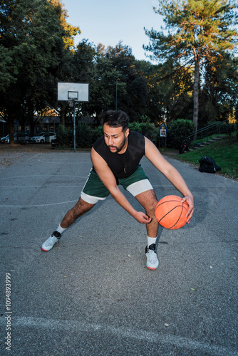 A man in a black tank top and green shorts carefully guards the basketball on an outdoor court surrounded by trees and lit by the setting sun. photo