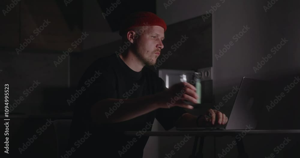 A man in black clothes is working at home in the dark using a laptop and sitting in the kitchen. A man drinks an invigorating drink, energy drink or coffee to stay awake and work all night. 4k footage A man in black clothes is working at home in the dark using a laptop and sitting in the kitchen. A man drinks an invigorating drink, energy drink or coffee to stay awake and work all night. 4k footage