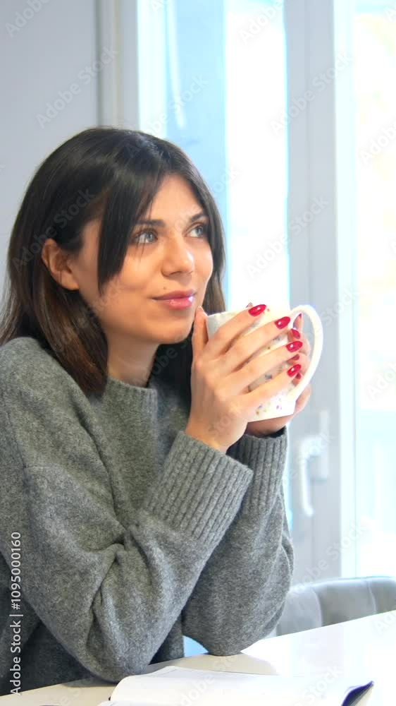A woman is drinking coffee while sitting at a table with a notebook. She is wearing a gray sweater and has red nails A woman is drinking coffee while sitting at a table with a notebook. She is wearing a gray sweater and has red nails