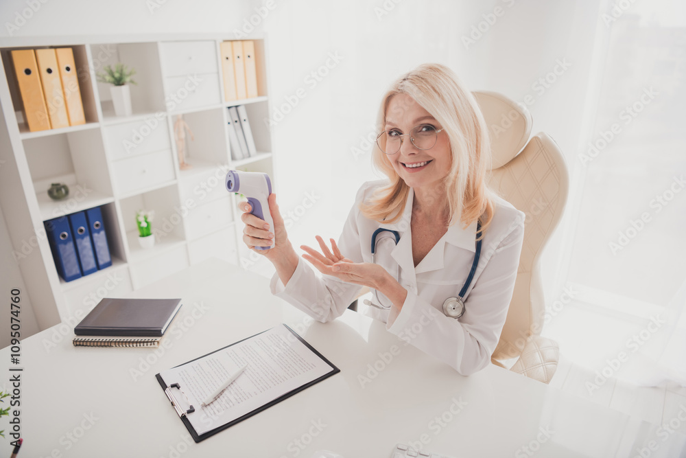 Smiling senior female doctor in a clinic office offering temperature check with digital thermometer Smiling senior female doctor in a clinic office offering temperature check with digital thermometer