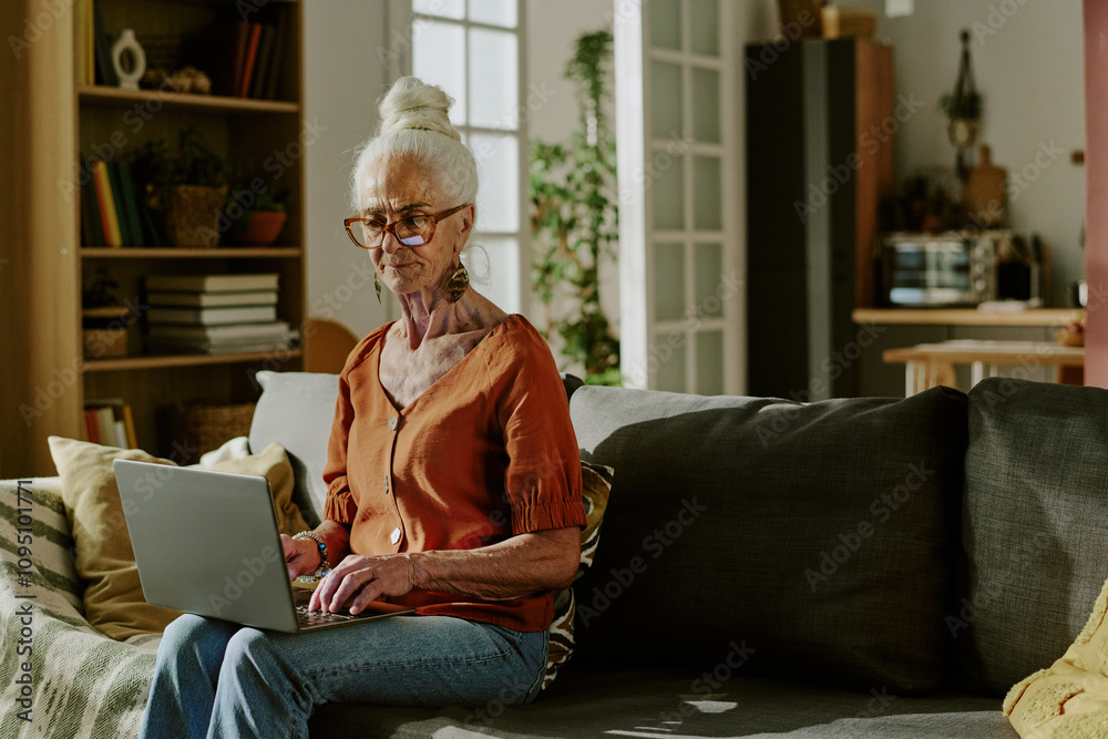 Senior woman with white hair sitting on a couch, using a laptop in a well-lit living room with modern decor, looking focused on her work Senior woman with white hair sitting on a couch, using a laptop in a well-lit living room with modern decor, looking focused on her work