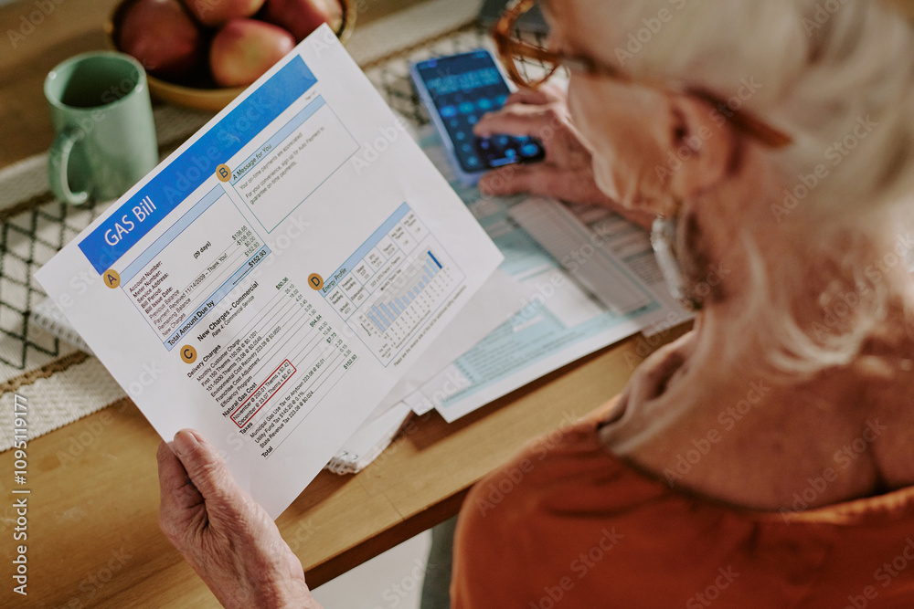 Obraz premium Senior woman examining gas bill and calculating expenses while sitting at dining table. Home setting includes papers, calculator, fruit bowl, and green cup