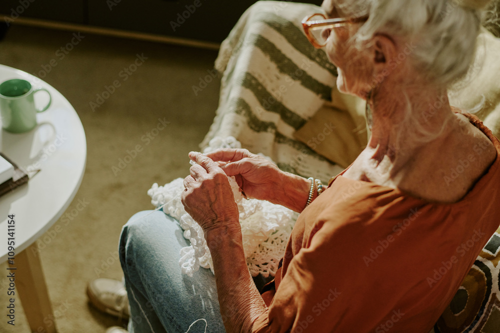Obraz premium Senior woman sitting indoors while crocheting with focused expression. Warm lighting providing cozy atmosphere as she works on handmade project