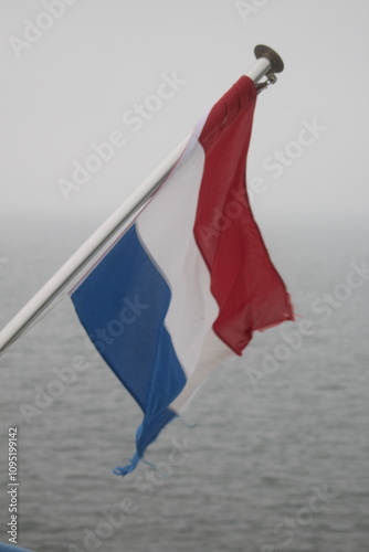 A Dutch flag, with its distinctive colors of red, white, and blue, flutters in the wind against a backdrop of a gray, overcast sky and a calm sea. The flag is attached to a flagpole on a boat