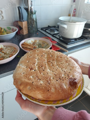 A freshly baked pita bread, still warm and soft, being held up by a hand. The bread is round and has a golden-brown crust with sesame seeds. In the background, there are bowls of soup and other dishes