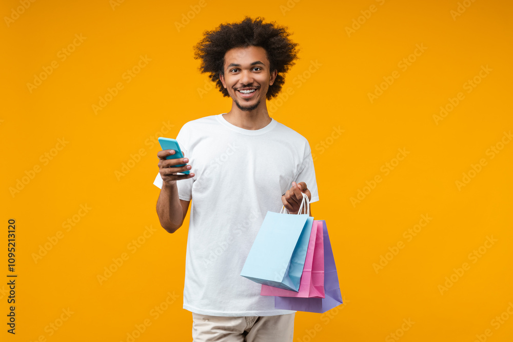 Portrait of attractive smiling young man customer posing in studio over bright colored orange yellow background with a bunch of shopping bags and smartphone in hands Portrait of attractive smiling young man customer posing in studio over bright colored orange yellow background with a bunch of shopping bags and smartphone in hands