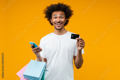 Portrait of joyful young man customer posing in studio with a bunch of shopping bags, credit card and smartphone in hands with amazed face expression, isolated over orange yellow background