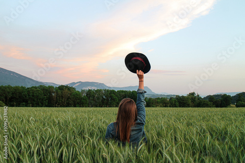 Sunset picture of a woman in a green wheat field raising a black hat to the sky. Peace of mind, freedom, and a deep connection with nature. Pastel sky, clouds, mountains, and forest in the background.