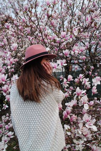 Portrait picture of a girl standing in front of a pink tree holding her face with her hand. Woman with a pink hat and a white winter jumper posing next to flowers in bloom. Long dark and blonde hair. 