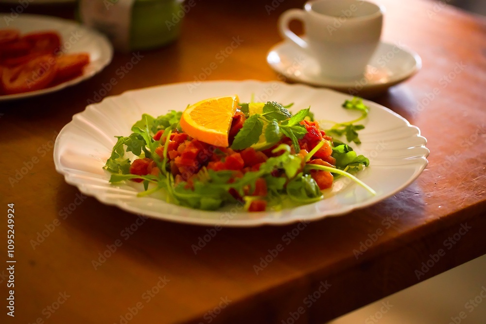 White plate with tuna tartare, a touch of arugula, orange, lime, and mint on a wooden table, surrounded by breakfast dishes. White plate with tuna tartare, a touch of arugula, orange, lime, and mint on a wooden table, surrounded by breakfast dishes.