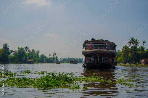 Small houses in a local village located next to Kerala's backwater on a bright sunny day and traditional Houseboat seen sailing through the river  Backwaters Alappuzha  , Kerala, India