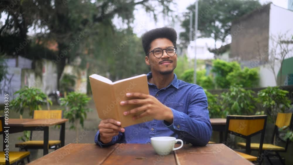 Young African American man in glasses reading book outdoors at a cozy cafe. Thoughtful man in casual denim shirt enjoying peaceful moment with coffee on a rustic wooden table. Young African American man in glasses reading book outdoors at a cozy cafe. Thoughtful man in casual denim shirt enjoying peaceful moment with coffee on a rustic wooden table.