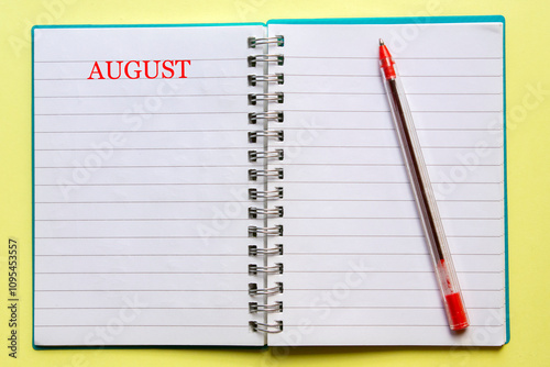 the word AUGUST written on an open notebook, with a red pen next to , on yellow background. Top view