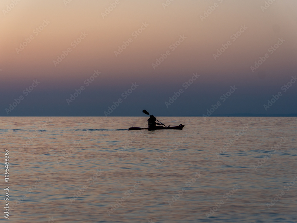 Naklejka premium A person paddles gently across tranquil waters as the sun sets, casting warm orange and soft blue tones across the horizon. The peaceful moment captures the essence of evening solitude.
