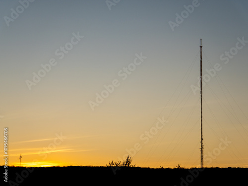 A tall communication tower stands silhouetted against the vibrant sunset sky. Warm colors blend as the day transitions to night in a peaceful, remote area.