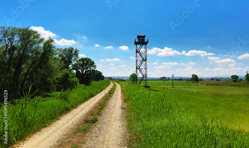 A rural watchtower stands tall beside a winding dirt road surrounded by lush green fields and trees under a vibrant blue sky with scattered white clouds. photo