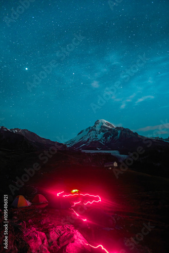 Under the blanket of stars, an otherworldly red light trail snakes through the rocky terrain. The Kazbegi mountain's silhouette