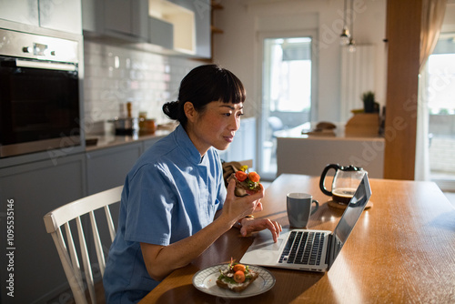 Asian woman eating healthy breakfast while working on laptop at home kitchen table
