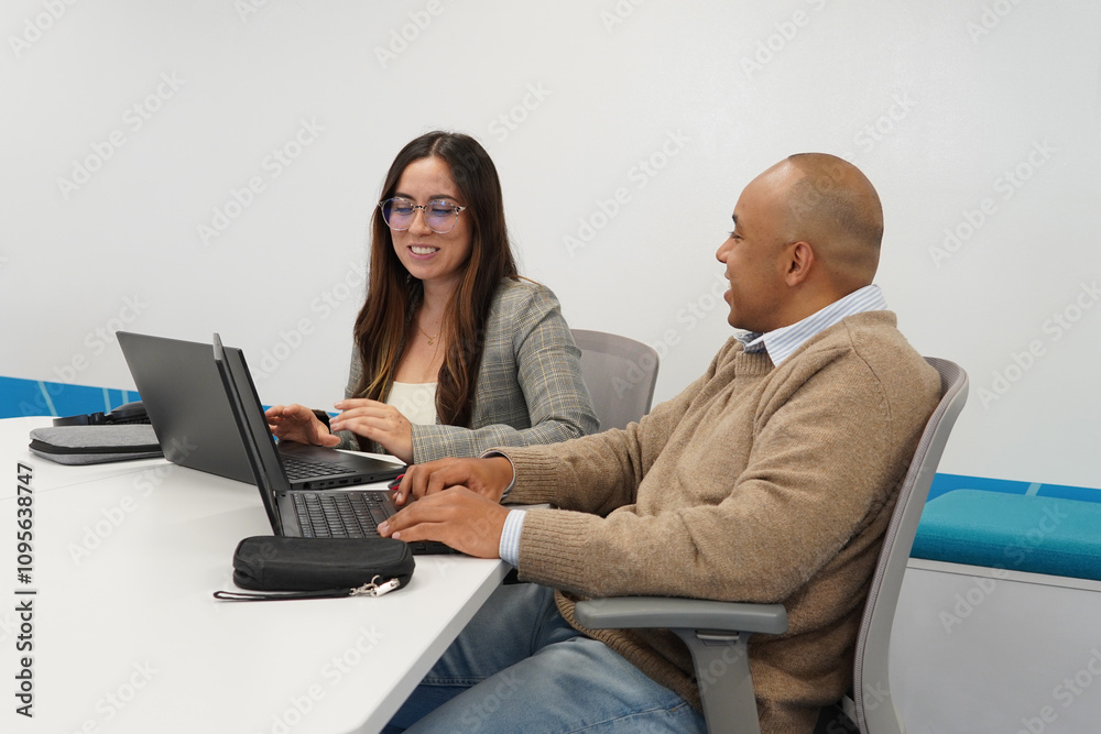 A couple of young coworkers sitting next to each other, both smiling. The woman has glasses and the man is bald. They are both in front of two laptops. A couple of young coworkers sitting next to each other, both smiling. The woman has glasses and the man is bald. They are both in front of two laptops.