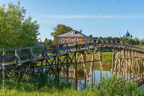 Russian traditional wooden house decorated with wood carvings on the background of a wooden pedestrian bridge over the Kamenka River on a sunny summer day, Suzdal, Vladimir region, Russia