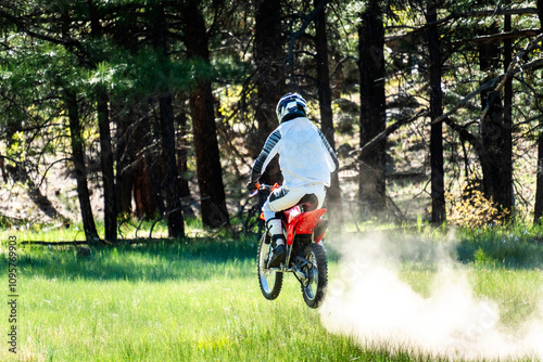 Motorcyclist jumps through grassy terrain in a forest during a sunny day