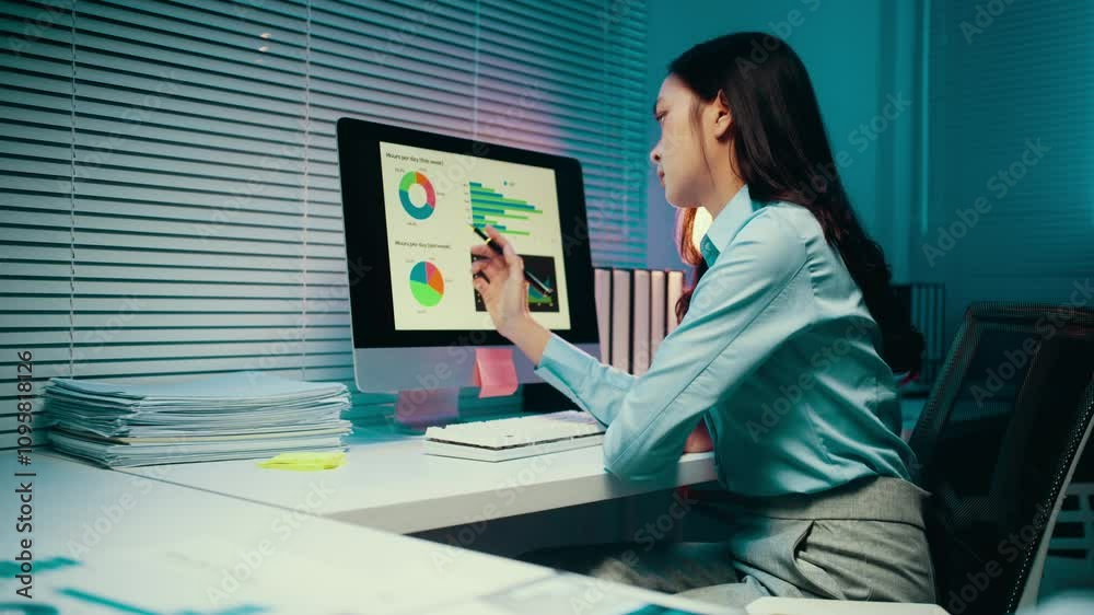 Businesswoman is analyzing financial data on a computer screen in her office. She is working late at night and is using a pen to point at the screen Businesswoman is analyzing financial data on a computer screen in her office. She is working late at night and is using a pen to point at the screen