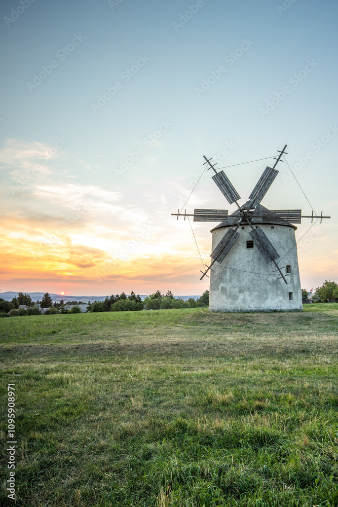 Windmill with wooden wings in a landscape setting. Fields, meadows and flowers appear in the sunset. Landscape shot in nature with a mill. historically untypical for the Balaton region in Hungary Windmill with wooden wings in a landscape setting. Fields, meadows and flowers appear in the sunset. Landscape shot in nature with a mill. historically untypical for the Balaton region in Hungary