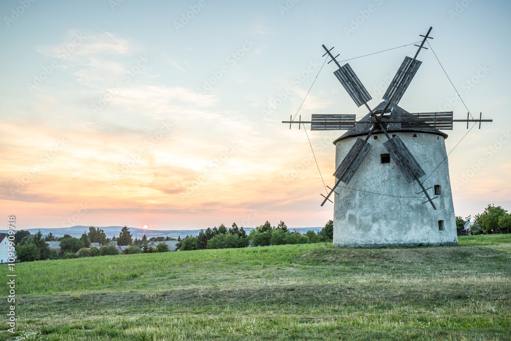 Windmill with wooden wings in a landscape setting. Fields, meadows and flowers appear in the sunset. Landscape shot in nature with a mill. historically untypical for the Balaton region in Hungary Windmill with wooden wings in a landscape setting. Fields, meadows and flowers appear in the sunset. Landscape shot in nature with a mill. historically untypical for the Balaton region in Hungary