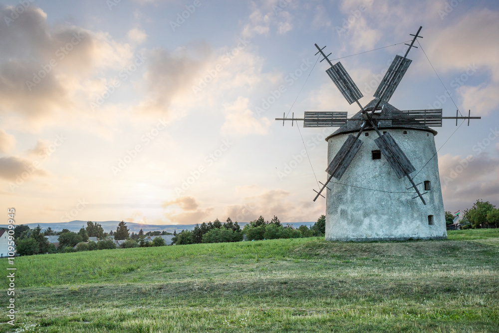 Windmill with wooden wings in a landscape setting. Fields, meadows and flowers appear in the sunset. Landscape shot in nature with a mill. historically untypical for the Balaton region in Hungary Windmill with wooden wings in a landscape setting. Fields, meadows and flowers appear in the sunset. Landscape shot in nature with a mill. historically untypical for the Balaton region in Hungary