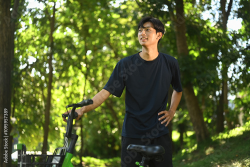 Smiling asian man standing near bike in serene park and enjoying the fresh air and natural surroundings