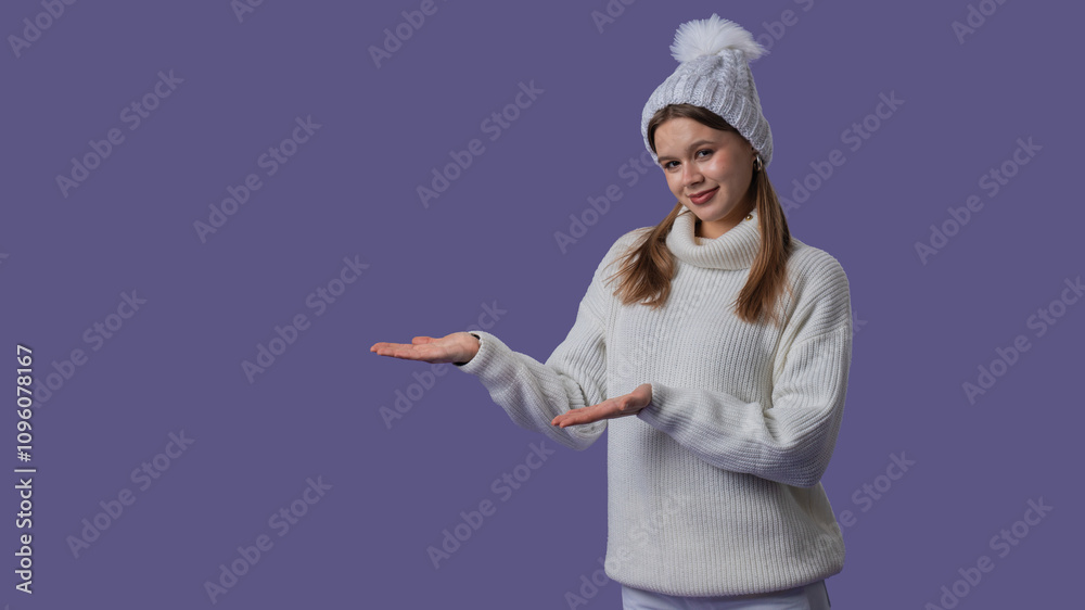 Cheerful young woman in white clothing and knitted hat smiles and shows at empty space with hands, isolated on purple studio background. Middle shot. Cheerful young woman in white clothing and knitted hat smiles and shows at empty space with hands, isolated on purple studio background. Middle shot.