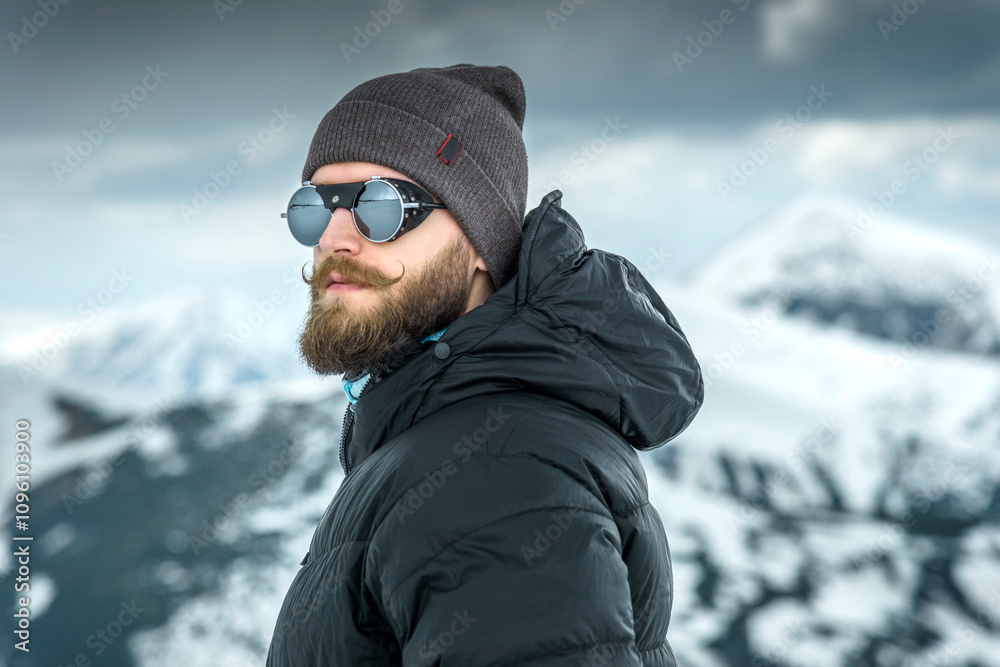 Portrait of a stylish man with a beard and curled mustache in the mountains with snow-covered peaks in the background and snow-laden clouds. Portrait of a stylish man with a beard and curled mustache in the mountains with snow-covered peaks in the background and snow-laden clouds.