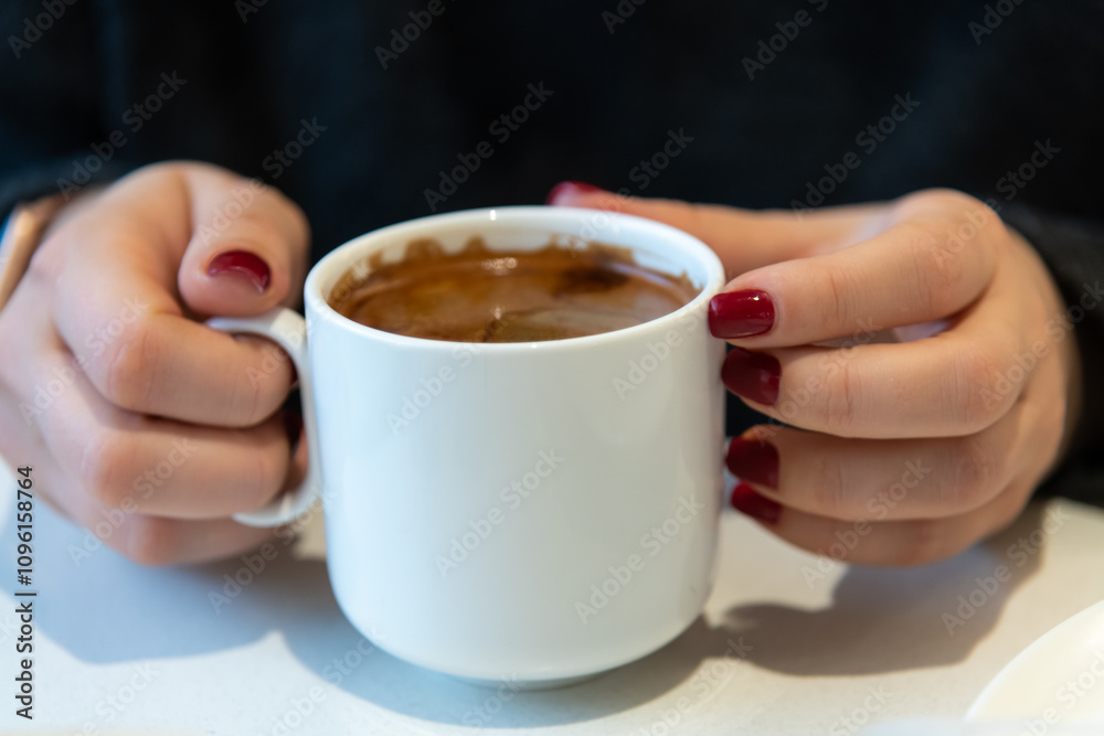 Close-up view of female hands with red fingernails holding white blank porcelain cup of americano coffee with brown crema by white table. Soft focus. Copy space. Hot drinks business theme. Close-up view of female hands with red fingernails holding white blank porcelain cup of americano coffee with brown crema by white table. Soft focus. Copy space. Hot drinks business theme.