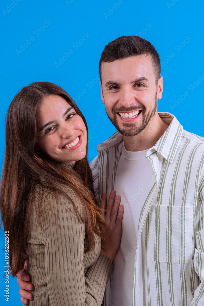 A joyful young couple embracing and smiling warmly at the camera against a vibrant blue background. Ideal image for love, relationships, happiness, or positive lifestyle concepts. A joyful young couple embracing and smiling warmly at the camera against a vibrant blue background. Ideal image for love, relationships, happiness, or positive lifestyle concepts.
