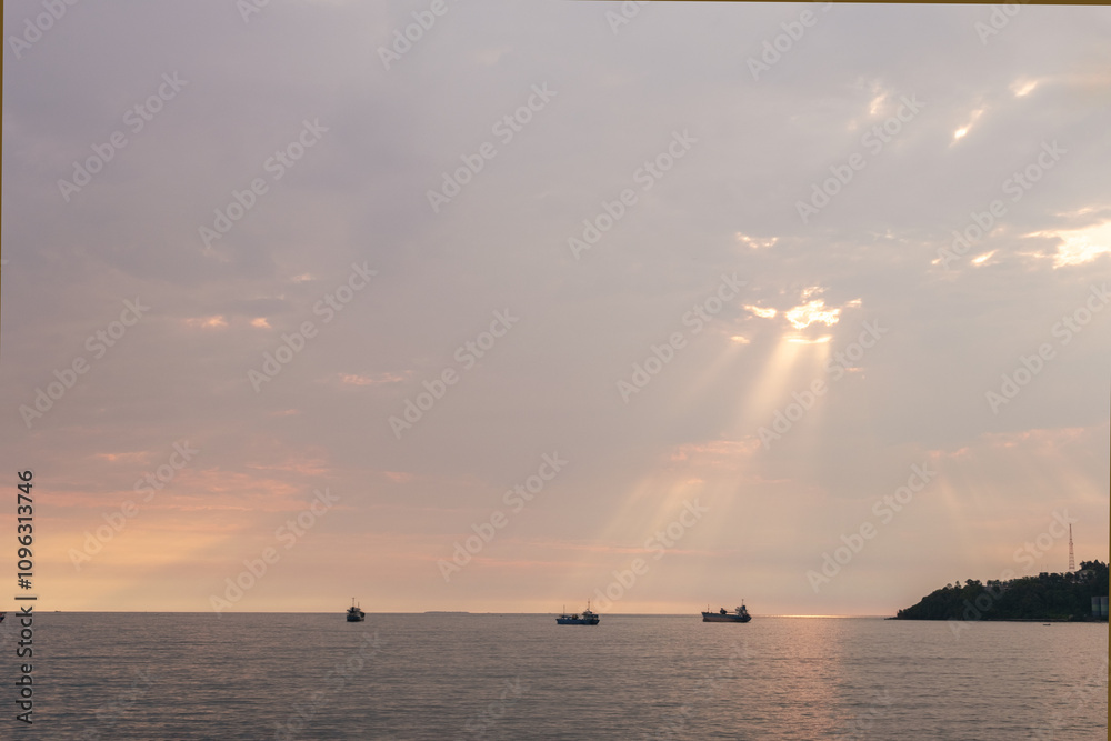 A cargo ship sails across calm waters under a cloudy sky as the afternoon sun shines through it, creating a warm golden glow as the sun sets. Perfect for nature, maritime and travel themes. A cargo ship sails across calm waters under a cloudy sky as the afternoon sun shines through it, creating a warm golden glow as the sun sets. Perfect for nature, maritime and travel themes.